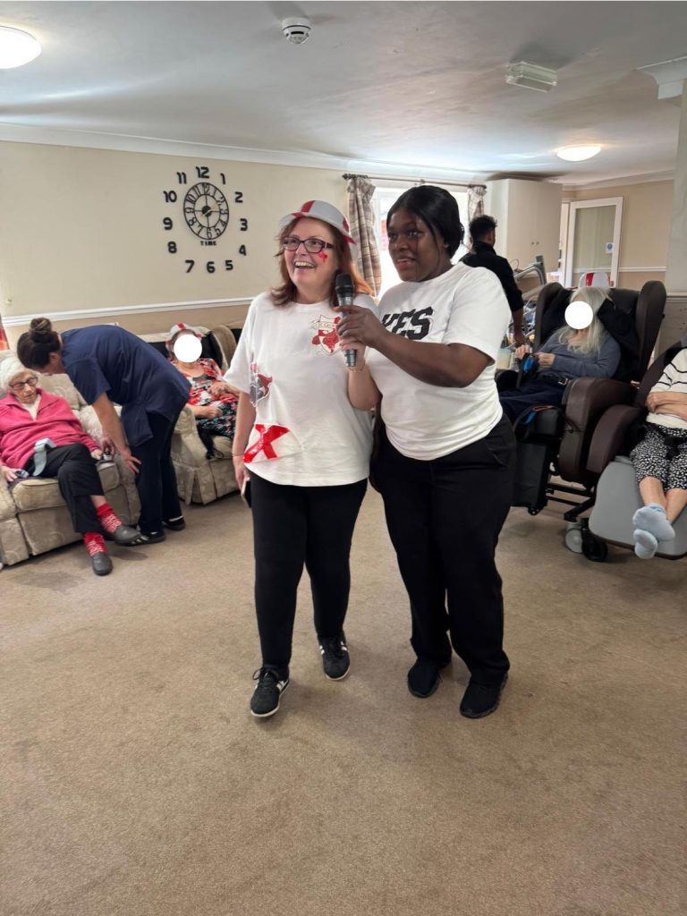 Two women smiling and singing into a microphone in a care home lounge.
