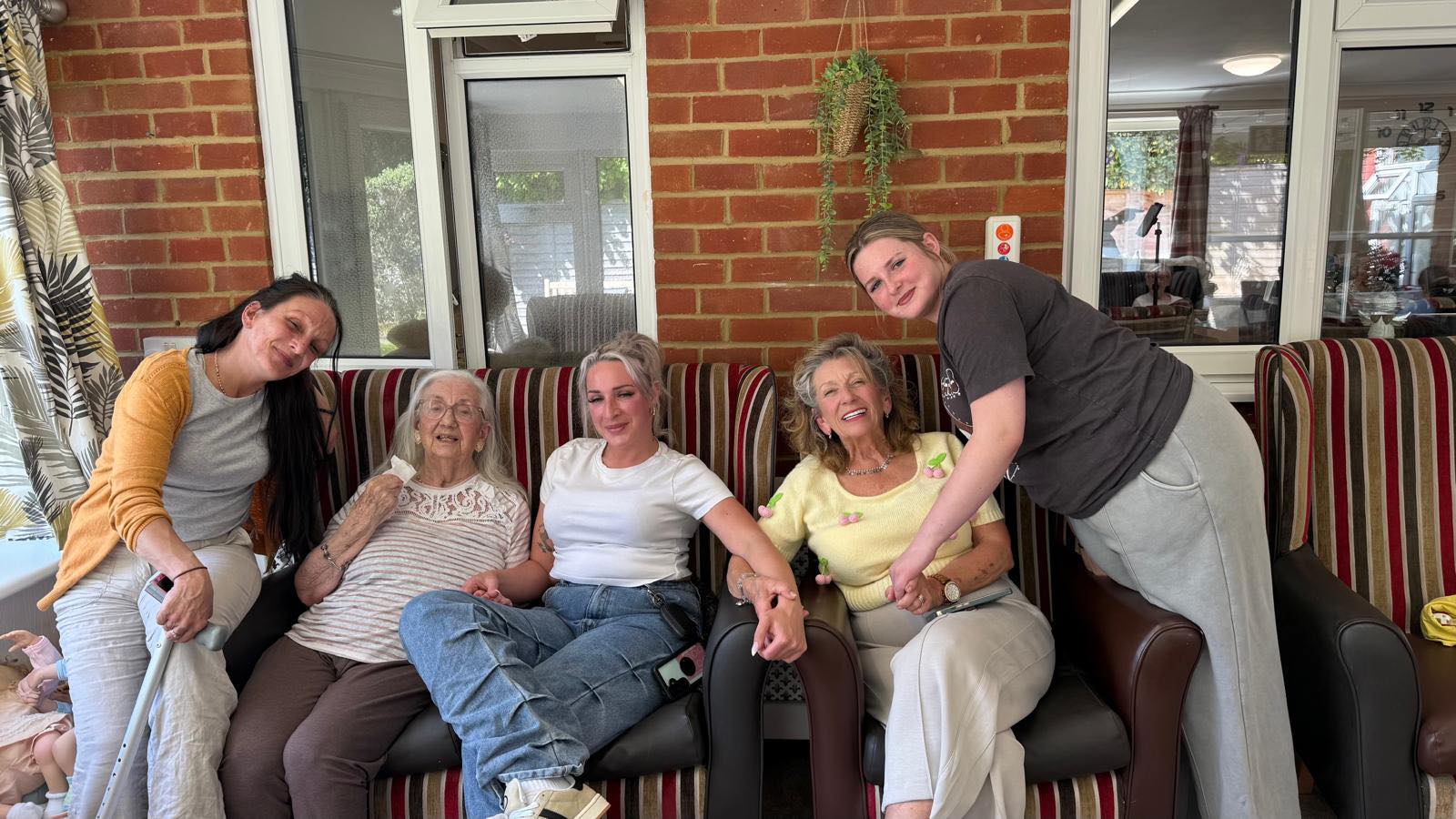 Five women of different ages sitting closely together on a sofa, smiling and holding hands in a warm, homely setting.