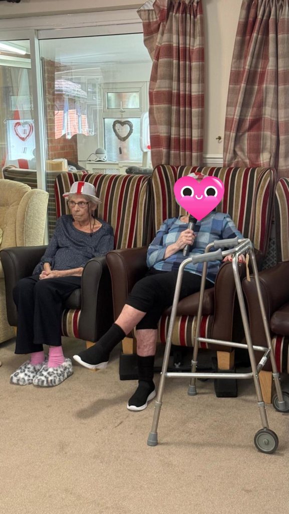 Two elderly women seated; one holding a microphone with a walker nearby.
