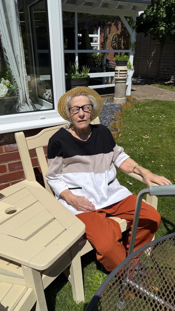Older woman sitting comfortably in the sunshine wearing a straw hat and glasses.
