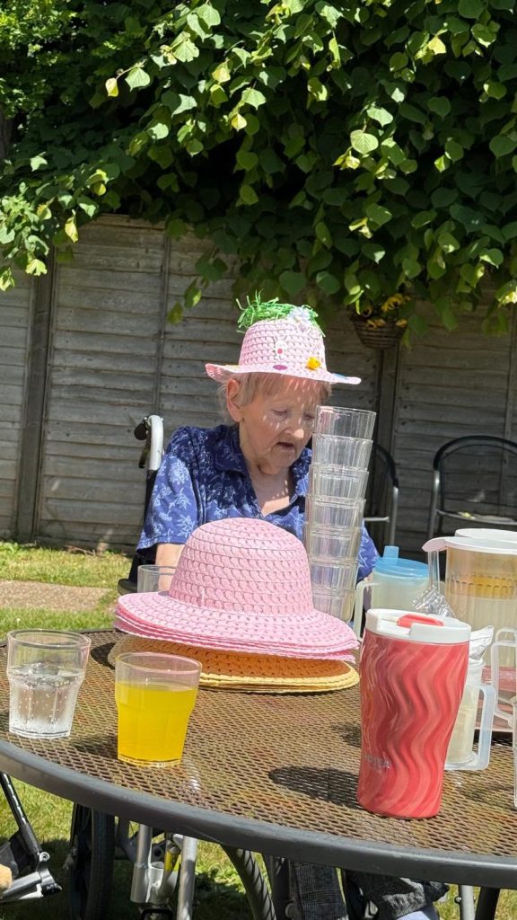 Elderly woman enjoying a sunny garden gathering with pink hats and drinks on the table.