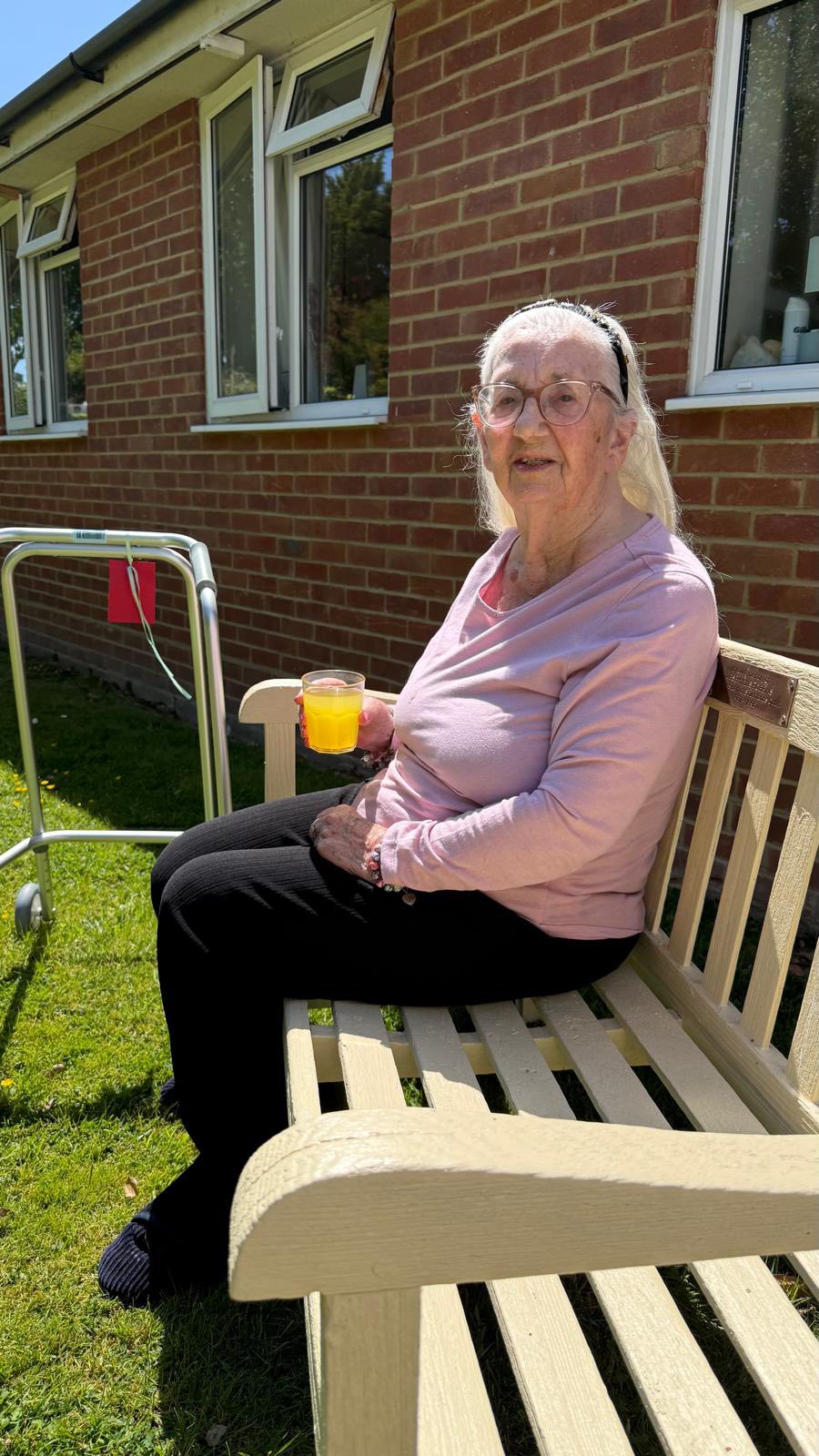 Elderly woman sitting on a garden bench enjoying a sunny day with a drink in hand.