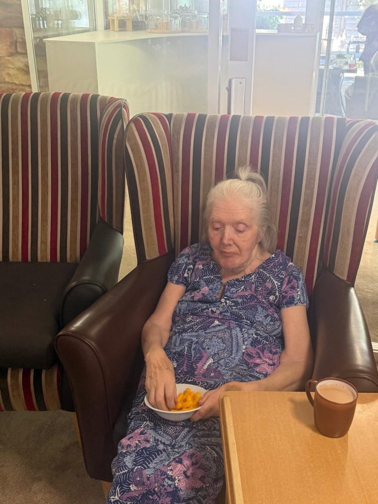 Elderly woman sitting in a high-backed chair, eating cheese puffs with a hot drink beside her.