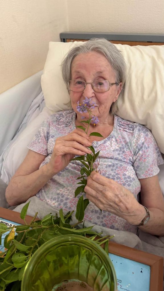 Elderly woman smiling as she smells a small bunch of purple flowers in bed.