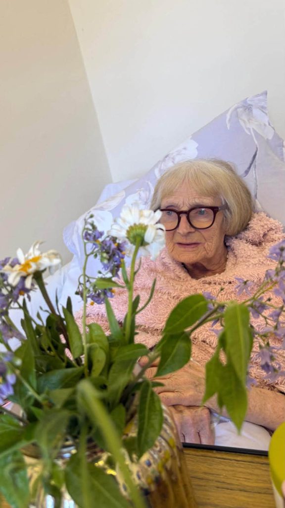 Smiling elderly woman sitting in bed with a colorful bouquet of fresh flowers in the foreground.