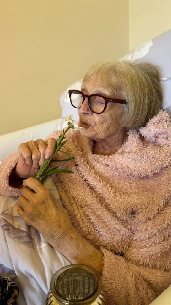 Elderly woman in a pink robe gently smelling a fresh white flower while relaxing in bed.