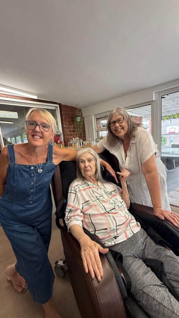 Three women pose together indoors, with one seated and two standing beside her.