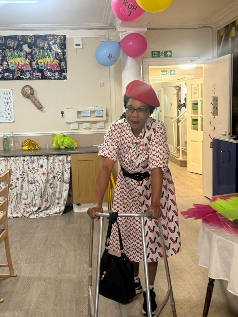 A woman wearing a red beret, patterned dress, and glasses stands with a walking frame in a decorated party room.