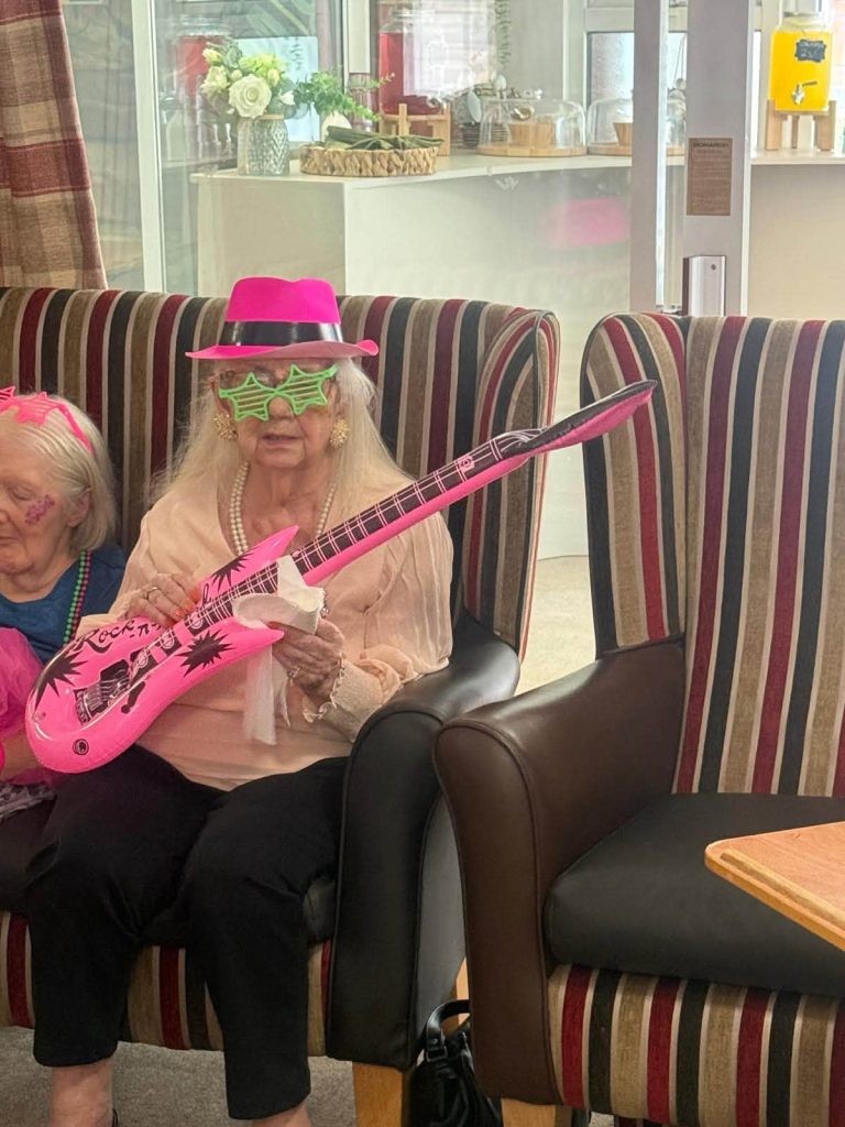 Care home resident wearing a pink hat and green glasses, holding a pink toy guitar at a themed party