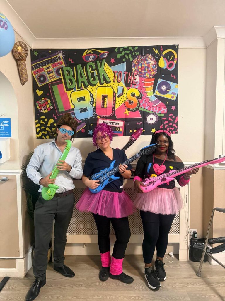 Care home staff posing with colourful toy guitars and 1980s decorations.