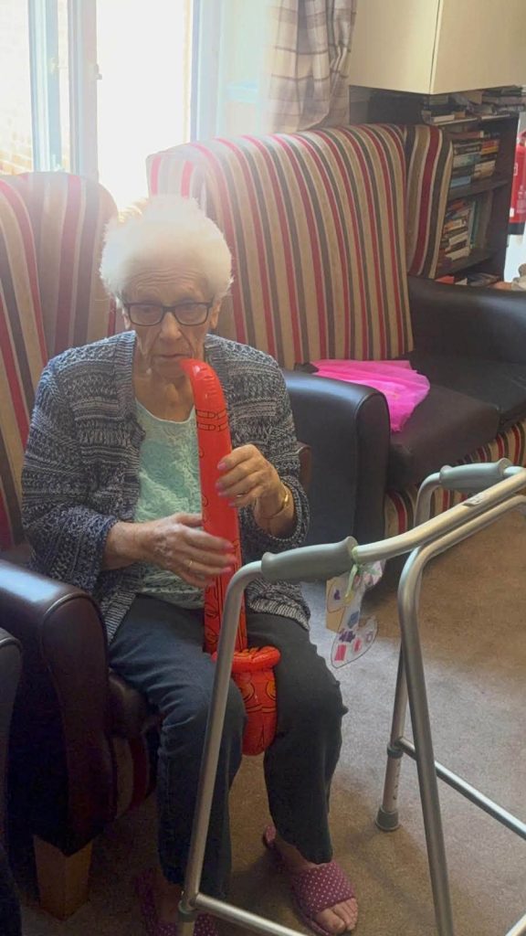 Elderly resident holding a colourful party prop while seated in a care home lounge.