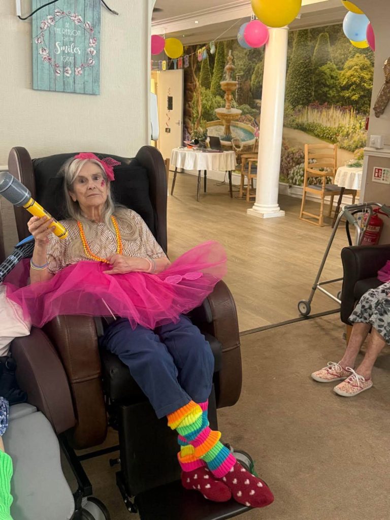 Resident dressed in a purple tutu and accessories, holding a blue inflatable guitar.