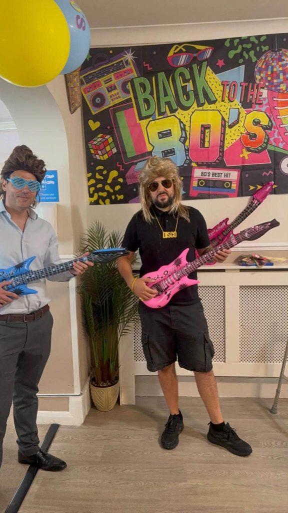 Staff members dressed in 1980s costumes with colourful toy guitars at a care home event.