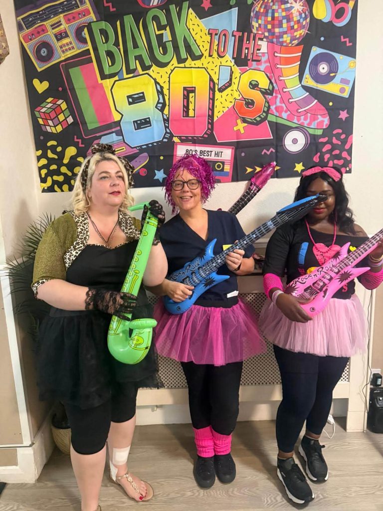 Staff members in retro costumes posing with toy guitars during a fun 1980s-themed care home party.