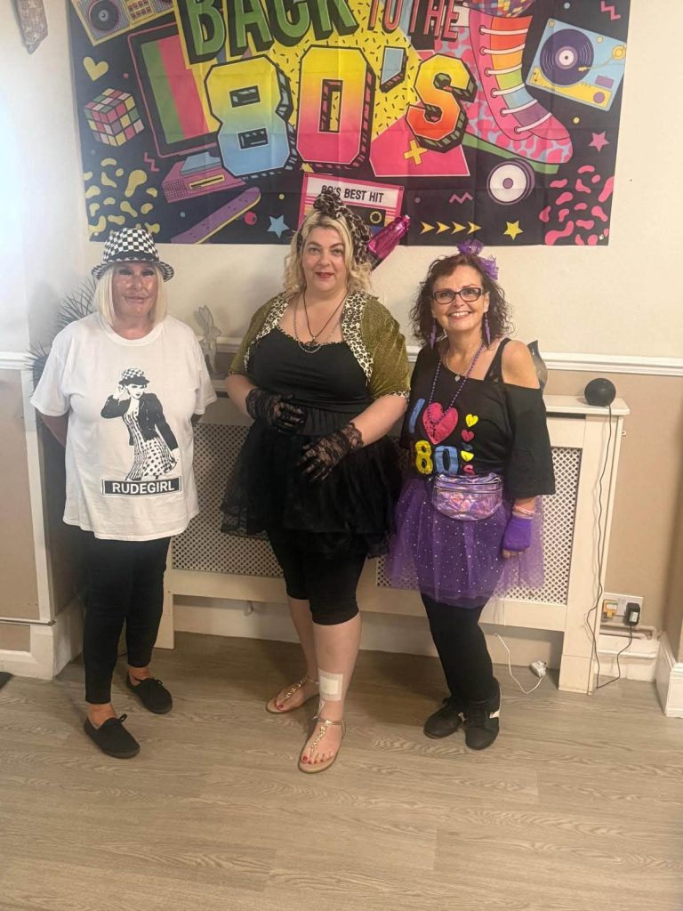 Three women pose together in 1980s-inspired outfits in front of a colorful โBack to the 80sโ party banner.