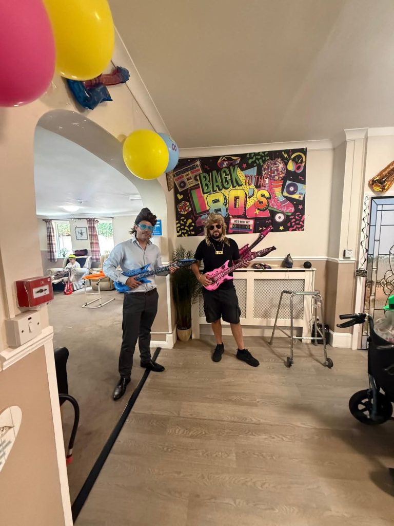 Two people dressed in retro 1980s outfits pose with colorful toy guitars at a themed party decorated with balloons and a โBack to the 80sโ banner.